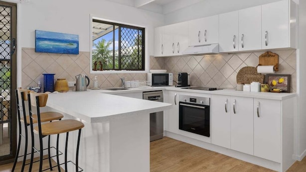 Una cocina moderna con armarios blancos, una isla grande con taburetes de bar, microondas, horno y ventana con vistas a palmeras.