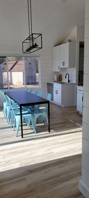 A modern kitchen with white cabinets, a dining area with a blue table and chairs, and a view of a house through the window.