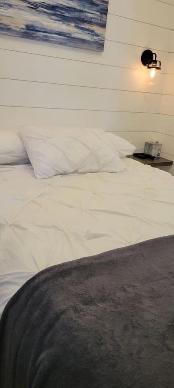Una cama con ropa de cama blanca, una manta oscura y una lámpara fijada a la pared.