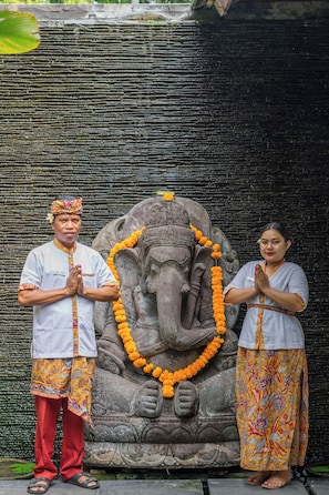 A stone statue of an elephant with intricate carvings, two people in traditional attire standing in front of it, and a textured wall behind them. - Villa Tigapuluh (Seminyak)