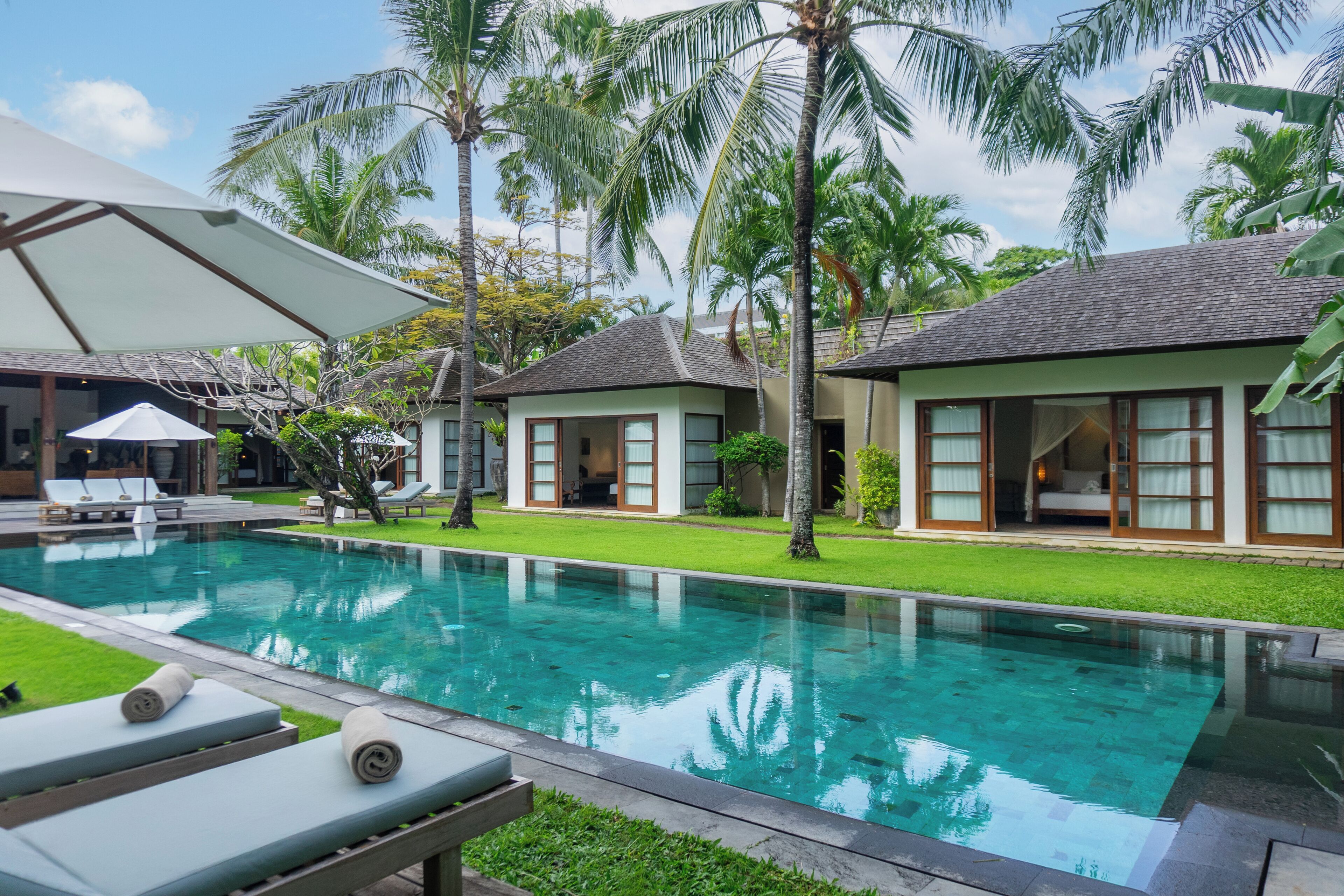 A poolside area with lounge chairs and a clear pool, surrounded by palm trees and a building with large windows.