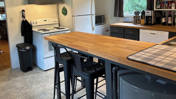 A compact kitchen with white cabinets, a wooden countertop, a stove, a refrigerator, a microwave, and a dishwasher. There are two bar stools and a trash can. - Wolf Creek Basecamp - Premier fishing! Walking distance to restaurants and bars (Wolf Creek)