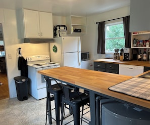 A compact kitchen with white cabinets, a wooden countertop, a stove, a refrigerator, a microwave, and a dishwasher. There are two bar stools and a trash can.