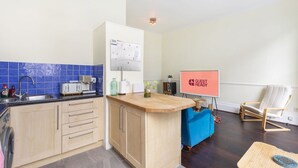 Ett kök med en diskho, brödrost och skåp. Där finns ett blått bord med en vit stol och en röd skylt med texten "KLART FÖR GÄSTER".