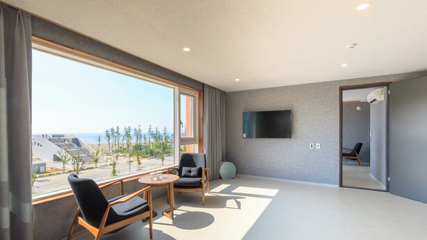 Une chambre d’hôtel moderne dotée d’une grande fenêtre donnant sur la plage et d’un balcon. Un téléviseur à écran plat est fixé au mur, et une table ronde est accompagnée de deux chaises.