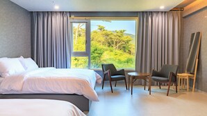 Une chambre d’hôtel moderne avec un grand lit, deux fauteuils, une petite table et une vue sur la verdure par la fenêtre.