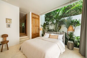 Een slaapkamer met een groot bed, een houten stoel, een kleine tafel en uitzicht op weelderige begroeiing door een raam.