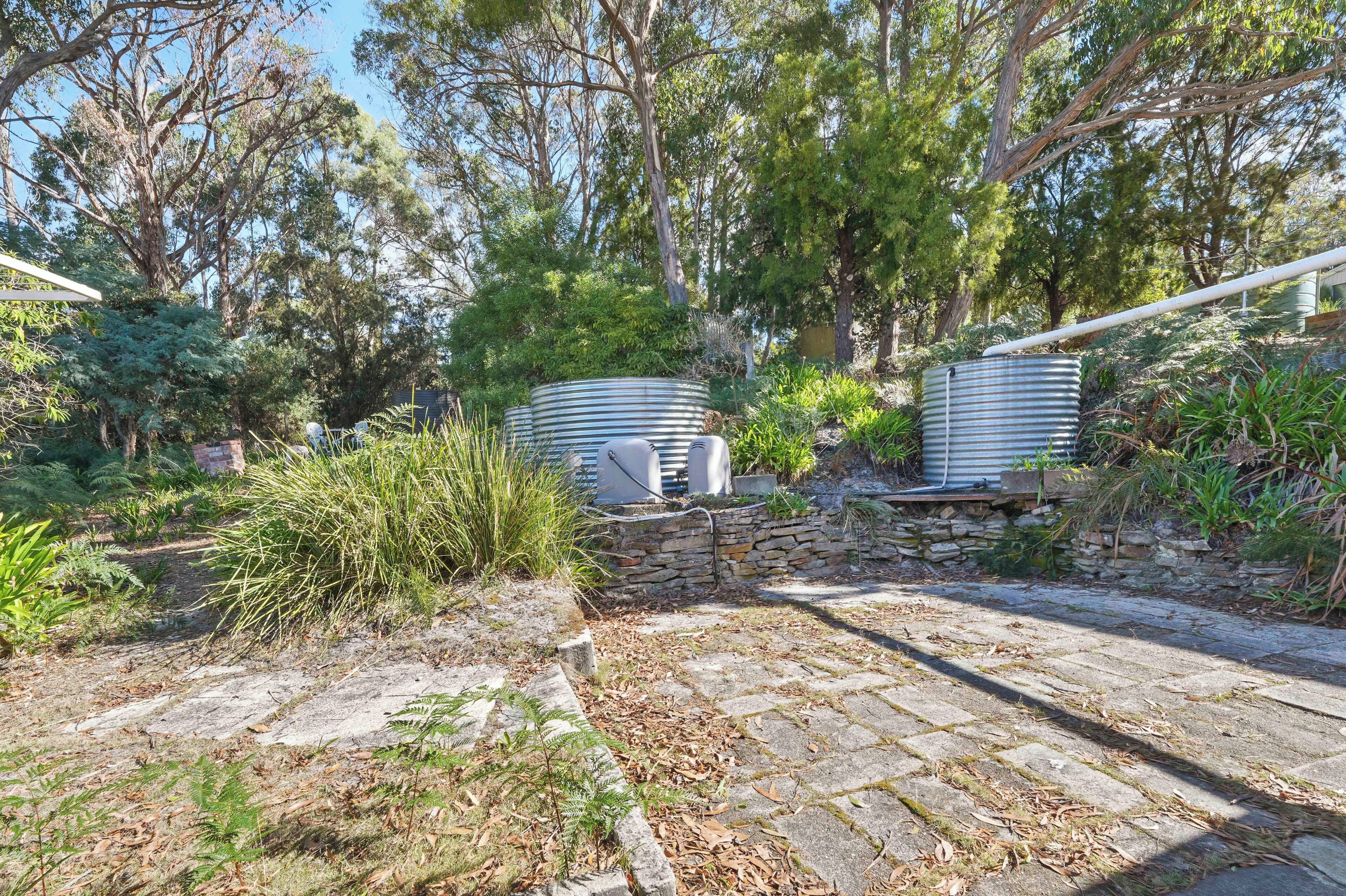 A stone pathway leading to a rustic outdoor area with metal water tanks, surrounded by dense greenery and trees.
