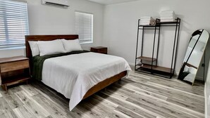 Ein Schlafzimmer mit einem hölzernen Bett, weißer Bettwäsche, einem Nachttisch aus Holz, einem Metallregal mit gefalteten Handtüchern und einem großen Spiegel.