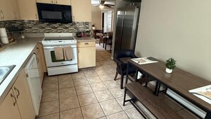 La cuisine est équipée d’un four à micro-ondes, d’un four et d’un réfrigérateur. On y trouve une table à manger avec des chaises et un banc. Le sol est carrelé et les murs sont peints dans une couleur neutre.