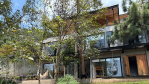 Una casa moderna de dos pisos con un amplio frente de vidrio, una terraza de madera y un jardín con diversas plantas y una hamaca.
