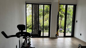 Una habitación amplia con una bicicleta estática, una cinta de correr y una colchoneta de yoga. Grandes ventanales ofrecen vistas a un jardín con árboles y plantas.