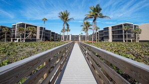 A wooden boardwalk leading to a modern building complex with multiple balconies and palm trees. - Charming 2-bedroom condo in fabulous Sanibel at Sundial (Sanibel)