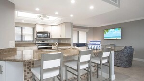 A modern kitchen with granite countertops, white cabinetry, and a built-in microwave. There is a bar area with four chairs and a television mounted on the wall. - Charming 2-bedroom condo in fabulous Sanibel at Sundial (Sanibel)