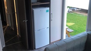 A room with a grey sofa, a white refrigerator, and a door leading to a balcony with a view of a grassy area.