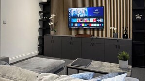 Un soggiorno moderno con una TV a schermo piatto montata su una parete in legno, divani grigi, un tavolino da caffè e un piccolo mobile contenitore con oggetti decorativi.