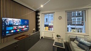 Un soggiorno moderno con una TV a schermo piatto montata a parete, un divano, un tavolino da caffè e una finestra con vista sugli edifici.