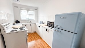 A compact kitchen with white cabinets, a white stove, a blue refrigerator, and a window providing natural light. - 2/7 Halloran Street, Wagga Wagga (Wagga Wagga)