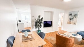 A modern living room with a wooden dining table, blue chairs, a brown ottoman, and a TV on a white stand. - 2/7 Halloran Street, Wagga Wagga (Wagga Wagga)