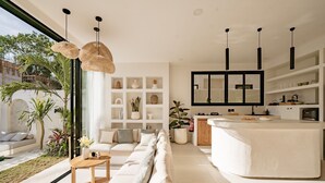 A modern living room with a large window, a sofa, a coffee table, and a kitchen area with a counter and cabinets. - Villa Yuni Uluwatu (Pecatu)
