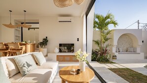 A modern living room with a sofa, a coffee table, and a TV. There is a dining area with a table and chairs, and a small garden with plants and a seating area with a stone wall. - Villa Yuni Uluwatu (Pecatu)