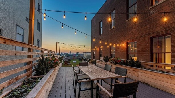 An outdoor patio with a wooden deck, a table with chairs, string lights, and a view of the cityscape. - Stylish Loft in St. Louis with Gym, Rooftop, Free Gated Parking, and Pool (St. Louis)