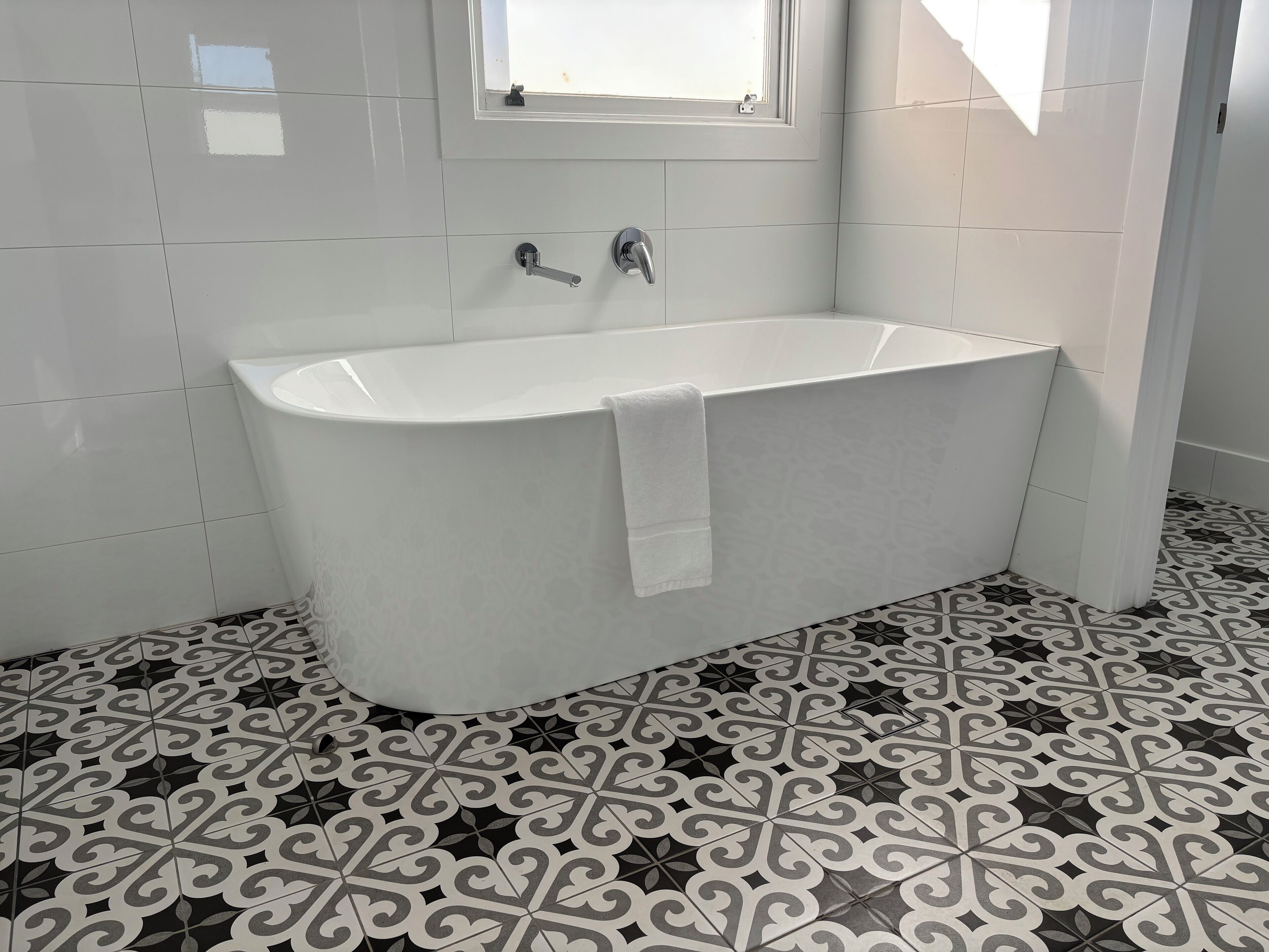 Een badkamer met een vrijstaand bad, een raam boven het bad en vloertegels met een patroon.