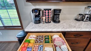 A kitchen with a marble countertop, coffee makers, and a drawer with various food items.