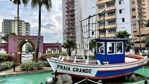 A boat named "Praia Grande" docked at a pool with a backdrop of multi-story buildings and palm trees. - Apartment 2 Blocks from Guilhermina Beach-Praia Grande SP! Near Boqueirão (Praia Grande)