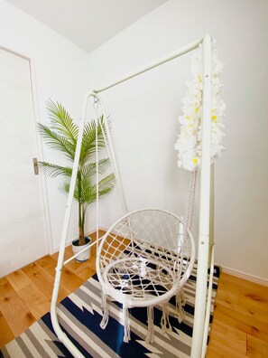 A white macrame chair hanging from a white metal frame, a potted palm plant, and a striped rug on a wooden floor. - New Onna | Cozy BBQ terrace, 8-min beach, snorkeling, SUP, sleeps 7, parking (恩納村)