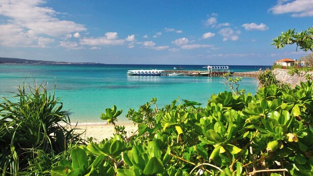 A coastal scene with a clear blue sea, a boat docked at a pier, and lush greenery in the foreground. - New Onna | Cozy BBQ terrace, 8-min beach, snorkeling, SUP, sleeps 7, parking (恩納村)