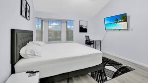 A modern bedroom with a large bed, a ceiling fan, a TV, and a view of the beach.