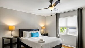 Una habitación de hotel con cama, dos mesitas de noche, ventilador de techo y ventana con cortinas.