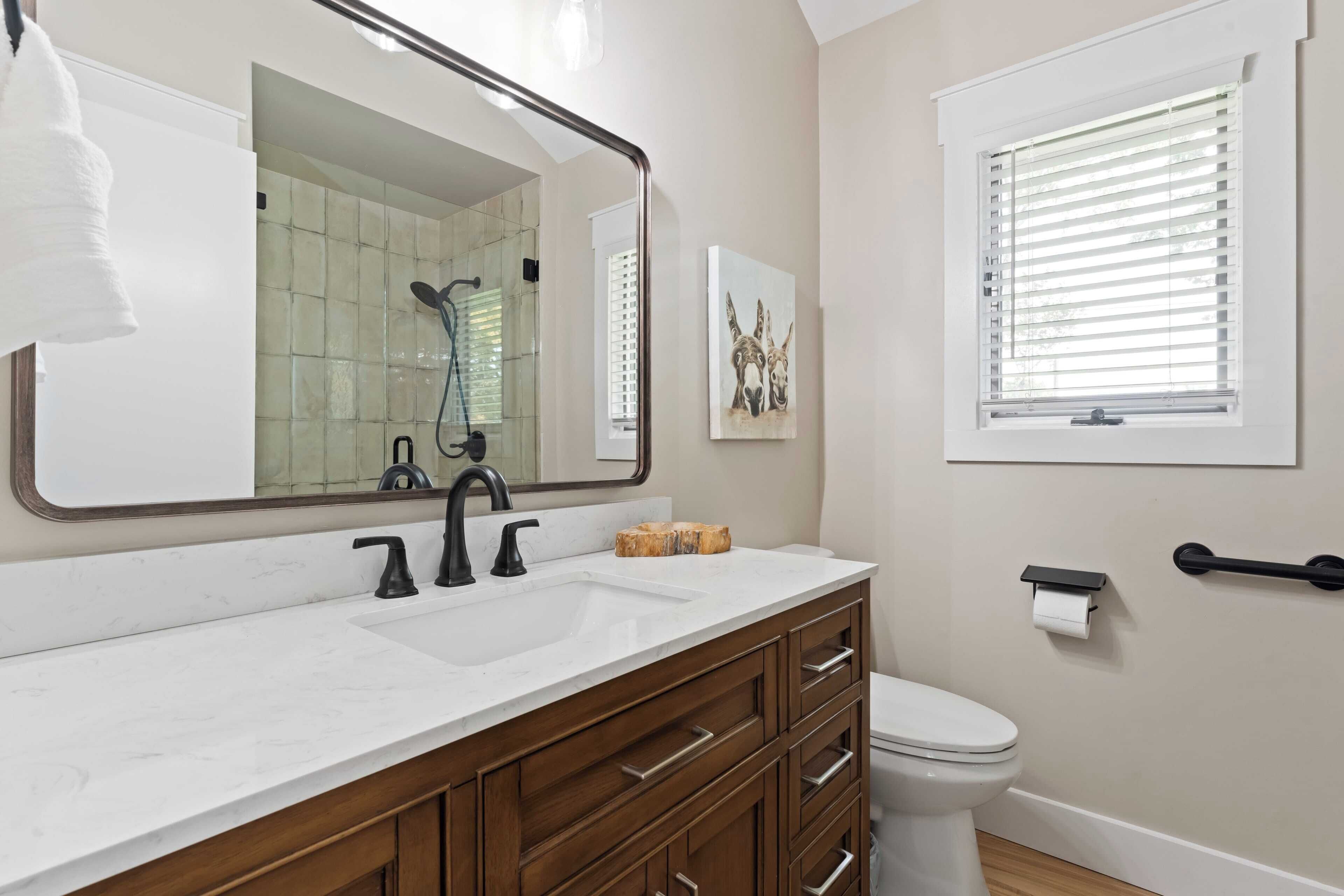 A bathroom with a white sink, two faucets, a mirror, a toilet, and a window with blinds.