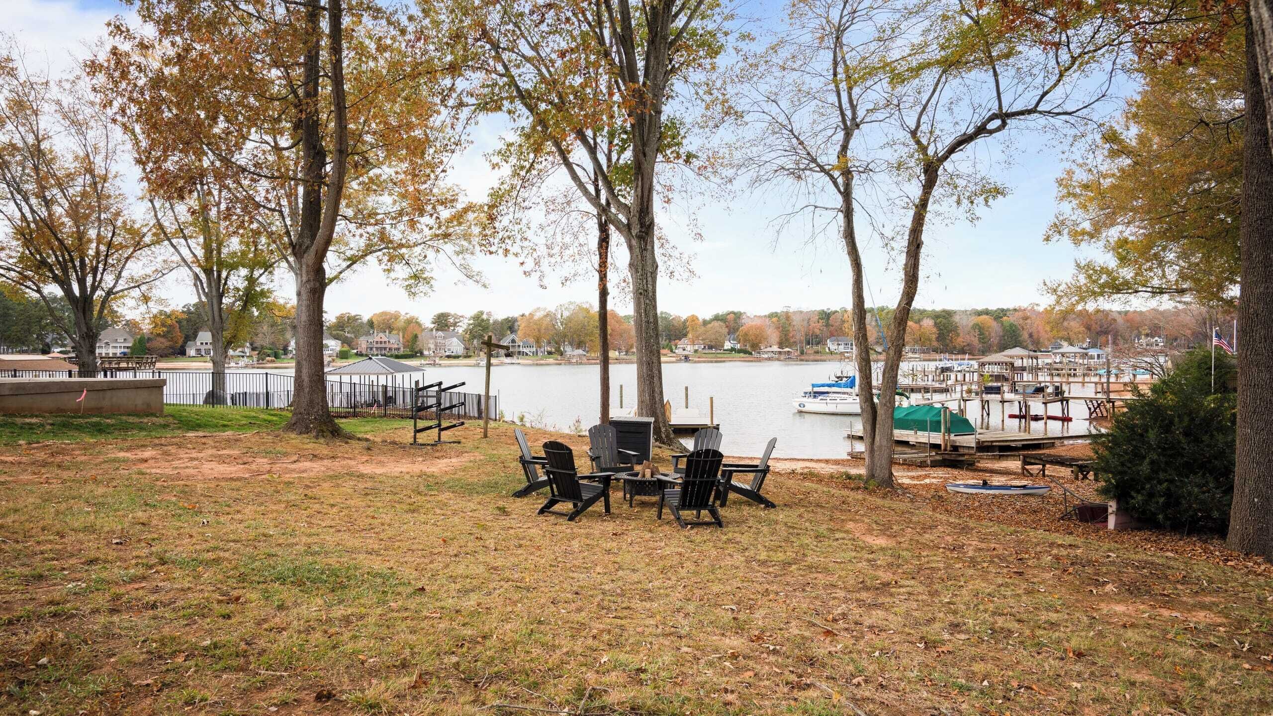 Un espace extérieur aménagé au bord du lac, avec des chaises Adirondack et un barbecue, entouré d’arbres et d’un quai où sont amarrés des bateaux.