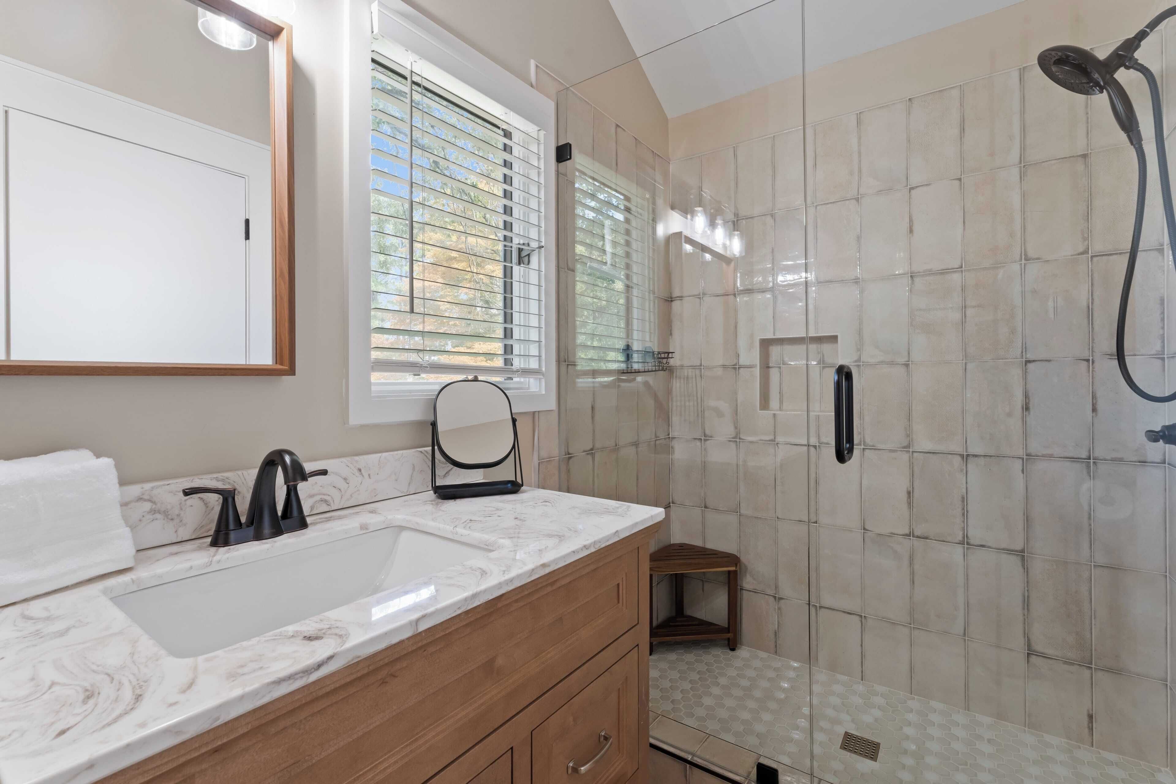 A bathroom with a marble sink, wooden vanity, a mirror, a shower with a handheld showerhead, and a window with blinds.