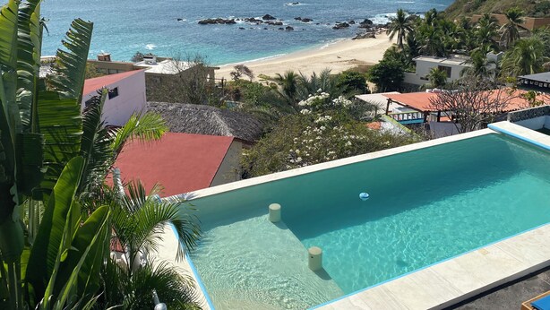 Una piscina con agua cristalina, rodeada de vegetación tropical y con vistas a una playa de costa rocosa.