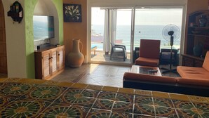 Una habitación con televisión, un florero, un ventilador y vista al mar a través de una puerta corrediza.