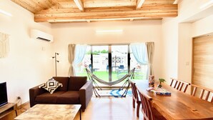 A living room with a sofa, coffee table, dining table, and chairs. There is a hammock, a television, and a large window with curtains.