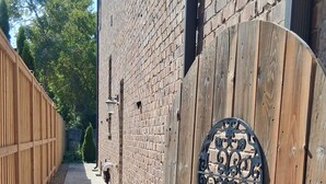 Un étroit sentier mène à un portail en bois orné d’une plaque métallique décorative, flanqué d’un mur de briques et d’une clôture en bois.