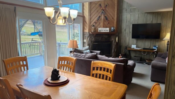 A dining area with a wooden table and chairs, a fireplace with a mounted television, and a view of the outdoors through large windows. - Charming 5BR House with Views of the Slopes, Full Kitchen, Patio, and Wifi (Beech Mountain)