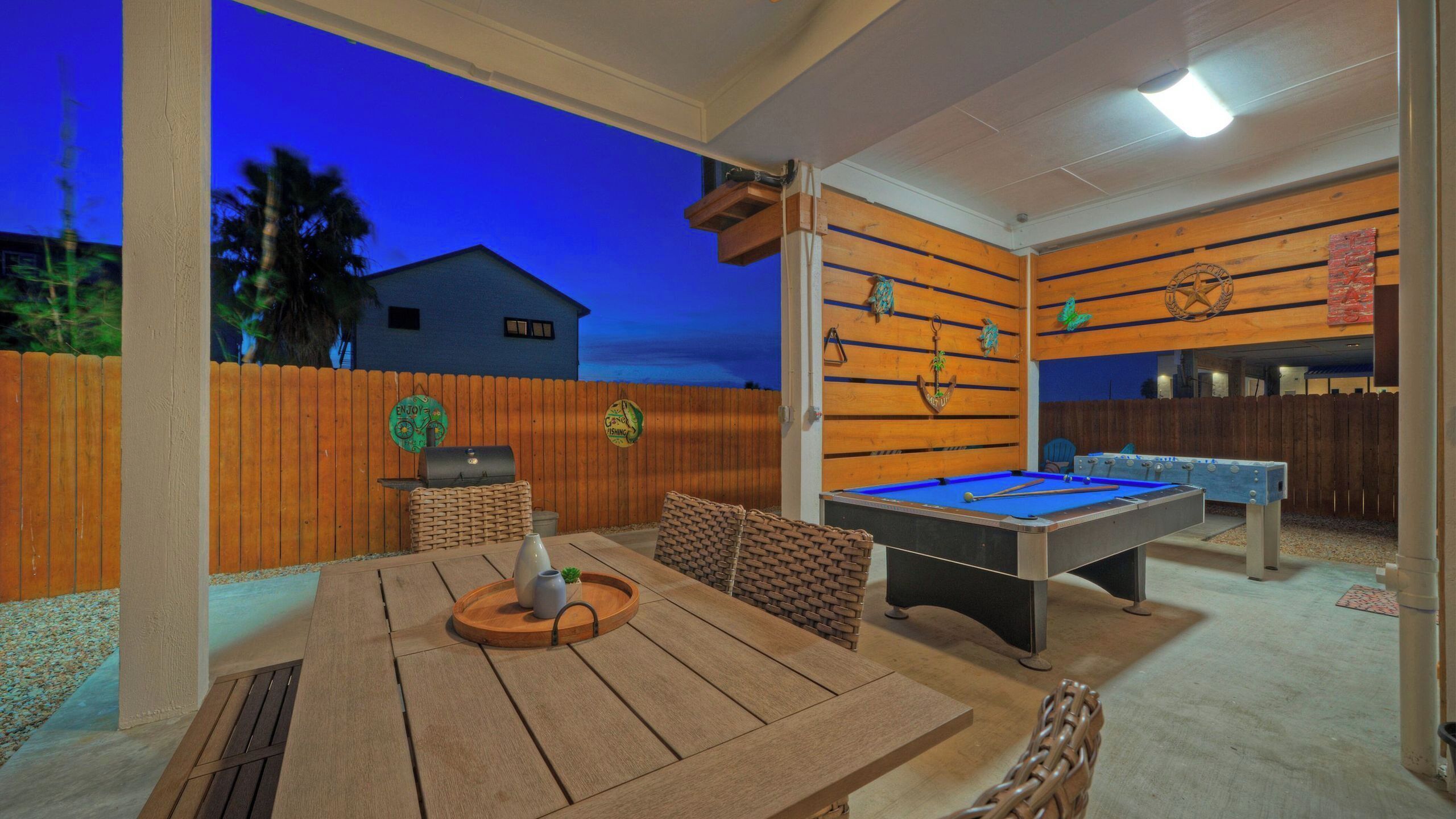 An outdoor pool table with a wooden table and chairs, a barbecue grill, and a wooden fence.