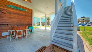 A covered outdoor area with a wooden bar and stools, a staircase, and a view of a house in the background.