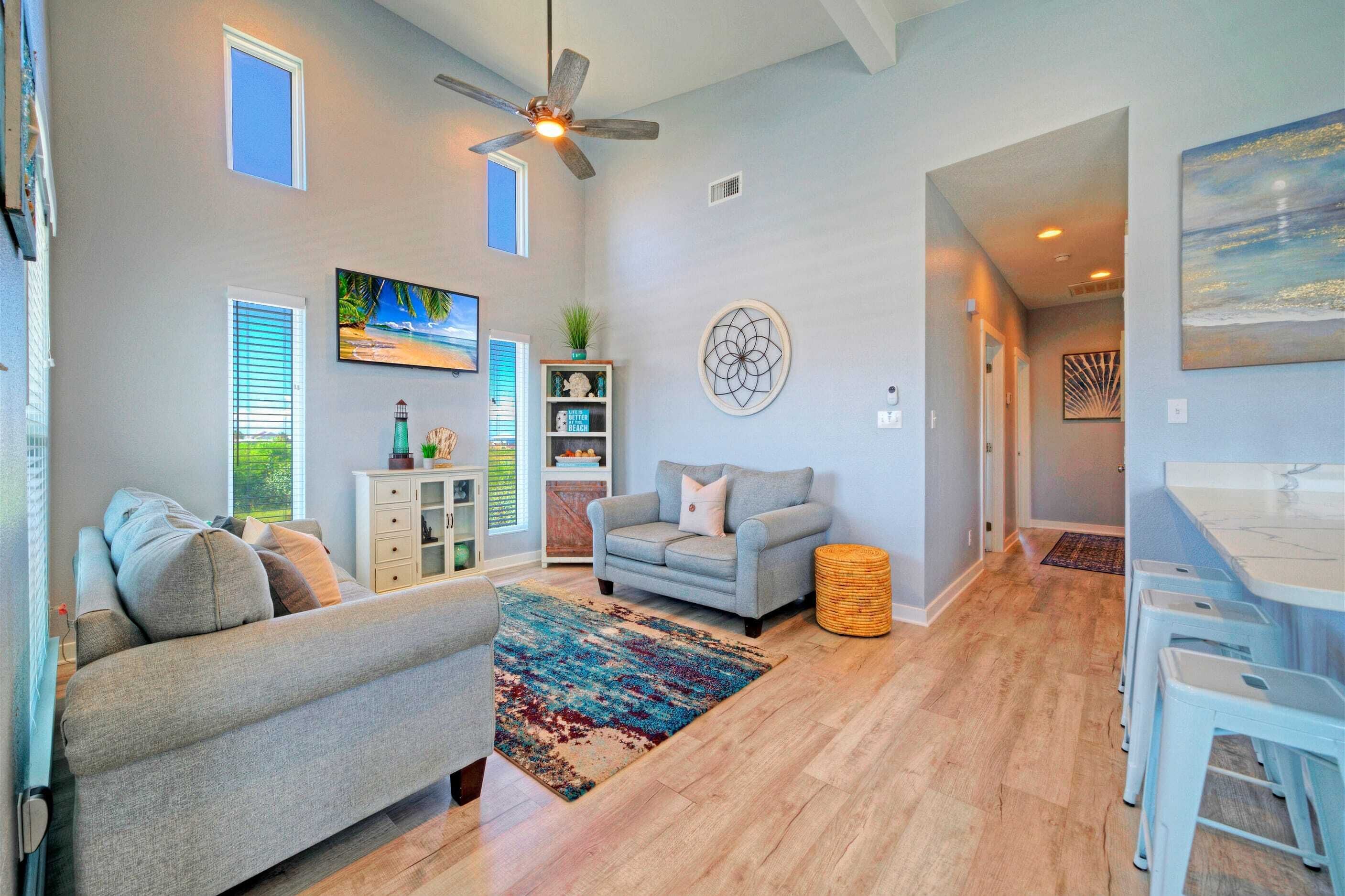 A spacious living room with a grey sofa, a coffee table, and a colorful rug. There is a ceiling fan, a painting, and a bookshelf with decorative items.