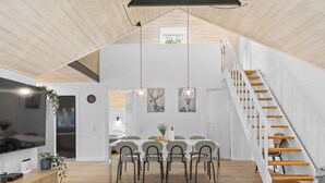 A spacious loft with a high ceiling, a dining area with a long table and chairs, a TV on the left, and a staircase on the right.