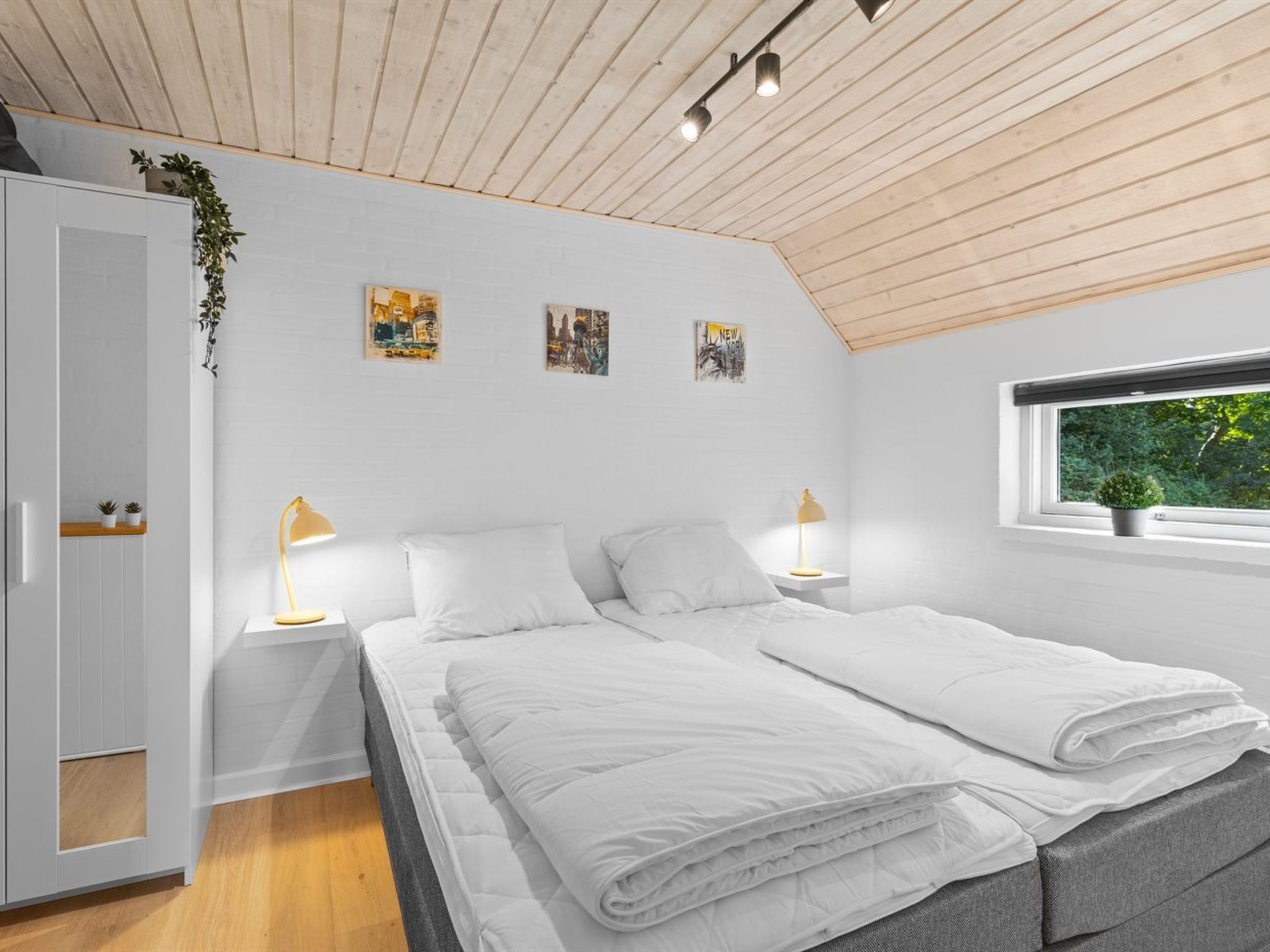 Ein modernes Schlafzimmer mit schrägen Wänden, weißen Wänden und einem großen Fenster mit Blick auf Grünflächen. Es gibt zwei Betten mit weißer Bettwäsche und zwei Nachttischlampen.