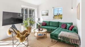 A living room with a green sofa, a round coffee table, a chair with a fur throw, a flat-screen TV, and a large window with a view.