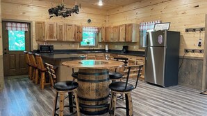 A wooden interior with a central island table, bar stools, a refrigerator, and wooden cabinets.