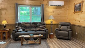 A cozy living room with a sofa, armchair, and coffee table, wooden walls, and a window with a view of greenery.