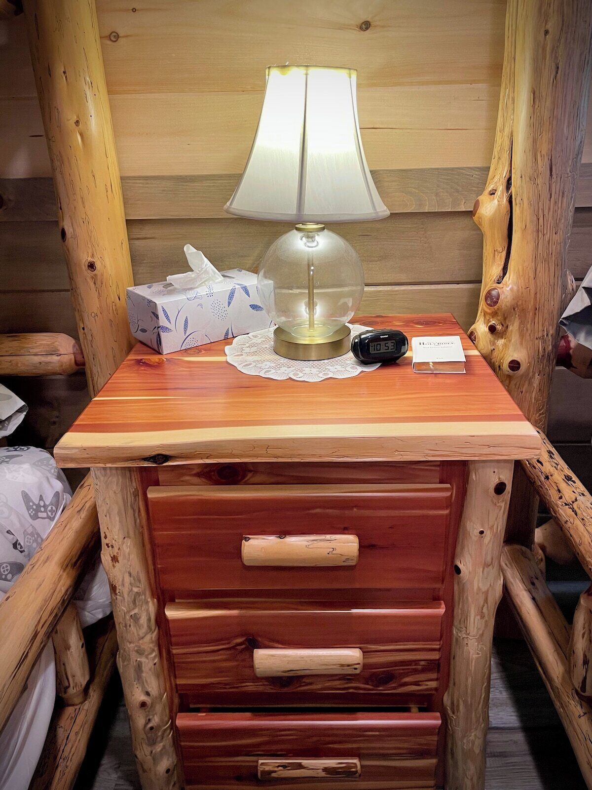 Ett litet träbord som står vid sängen, med en lampa, en näsduksask, en digital klocka och ett litet kort på.